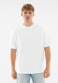 Biała bawełniana koszulka z krótkim rękawem, okrągłym dekoltem, luźnym krojem i minimalistycznym wzornictwem, z małym logo na obszarze klatki piersiowej.