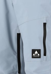 Ljusblå vattentät jacka med två svarta dragkedjefickor på sidorna och en fyrkantig logotyp med vit bergsdesign. Slät textur.