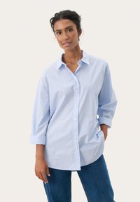 Camicia oversize a righe blu e bianche con bottoni frontali, maniche lunghe arrotolate e una texture in tessuto morbido. Indossata con jeans di denim.