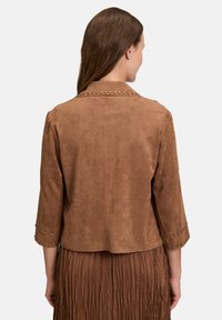 Veste en daim marron avec des manches trois-quarts, des accents en clous le long du col et des poignets, une texture douce et une silhouette ajustée.