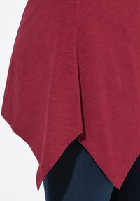 Bordeauxfarbenes Top mit abgerundetem Saum und spitzen Kanten, aus einem weichen, strukturierten Stoff, kombiniert mit dunkelblauen Hosen im Bild.