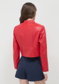 Rote, taillierte Lederjacke mit glatter Textur, strukturierten Schultern und einem subtilen Glanz. Kombiniert mit marineblauen Shorts für einen Kontrast.