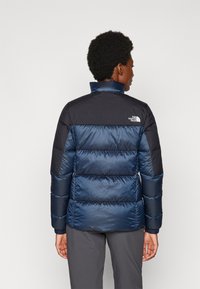 Námornícky modrá puffa bunda s kontrastnými čiernymi plecami, horizontálnym prešívaným dizajnom, vysokým golierom a malým logom na hornom chrbte.
