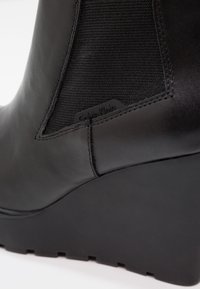 Bota de tobillo de cuero negro con paneles laterales de tela, suela con cuña, detalles cosidos y una pequeña etiqueta con el logo en el lado exterior.