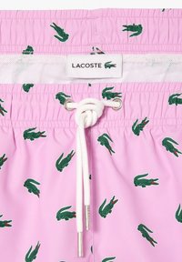 Shorts da bagno rosa con motivi di coccodrillo verdi. Caratterizzati da un laccio bianco con estremità metalliche e un'etichetta con marchio in vita.