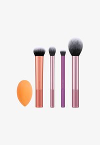 Cinq outils de maquillage alignés : une éponge à fondre orange et quatre pinceaux de tailles différentes avec des poils noirs et blancs et des manches métalliques.