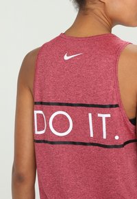 Punainen hihaton urheilutoppi, jossa on valkoinen "DO IT." -teksti ja musta raita. Suunniteltu hengittävästä kankaasta ja takana on Nike-logo.
