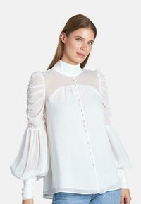 Blusa blanca con cuello alto, panel frontal transparente y mangas abullonadas. Presenta frente abotonado y tejido texturizado.