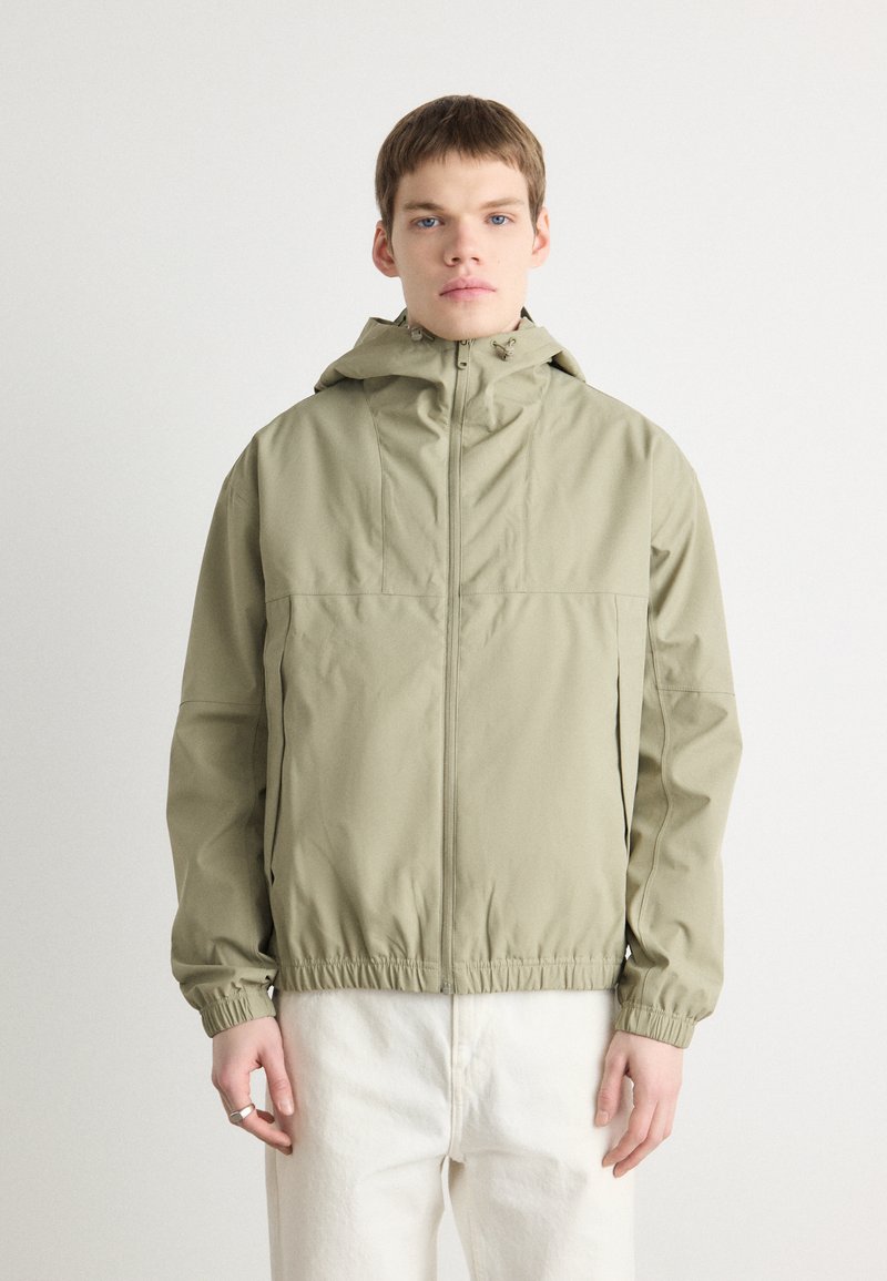 Light olive green jacket with a hood, featuring a zippered front, elastic cuffs, and a relaxed fit. Material appears smooth and water-resistant.