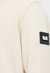 Beige sweatshirt featuring a textured fabric. Left sleeve displays a black rectangular patch with white text "WEEKEND OFFENDER."
