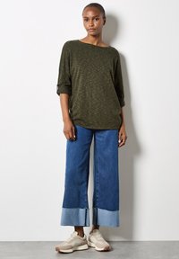 Olive green, textured top with a wide neckline and rolled sleeves. Blue wide-leg denim pants with frayed hem and light blue accent. Beige sneakers.