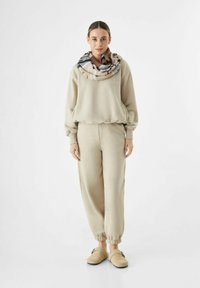 Sweatshirt beige en polaire et joggers assortis avec des poignets élastiques. Une écharpe à motifs drape autour du cou. Des chaussures souples à enfiler complètent l'ensemble.