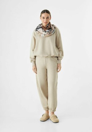 Beiger, fleeceartiger Sweatshirt und passende Jogginghosen mit elastischen Bündchen. Ein gemusterter Schal liegt um den Hals. Weiche Slipper runden das Outfit ab.