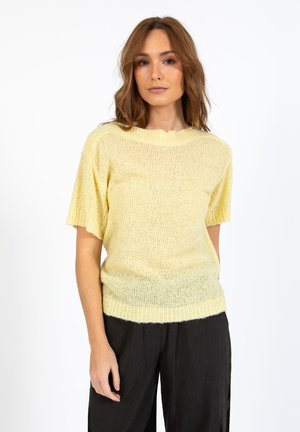 Mujer con el pelo ondulado hasta los hombros, que lleva un suéter corto de punto de color amarillo claro y pantalones negros holgados sobre un fondo blanco.
