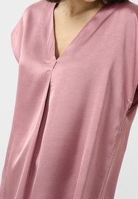 Haut en satin rose clair avec un col en V, des manches courtes et un pli central. Le tissu présente une texture lisse et un léger éclat.