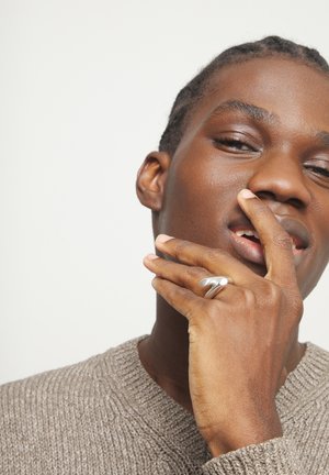 Primer plano de una persona con el cabello trenzado tocándose la cara, que lleva un suéter beige con textura y un anillo plateado en el dedo.
