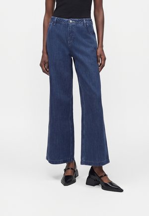 Persoană purtând blugi largi de culoare albastru închis, top negru fără mâneci și pantofi negri Mary Jane cu toc jos bloc, stând în fața unui fundal deschis.