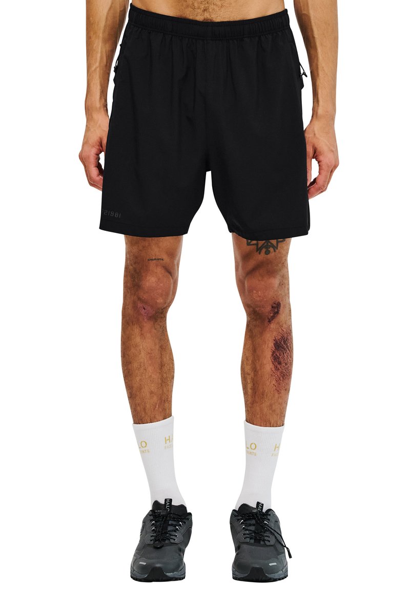Schwarze Sportshorts mit einem elastischen Bund, leichtem Material, Seitentaschen und einer Länge, die bis zur Mitte des Oberschenkels reicht. Getragen mit weißen Socken und schwarzen Sneakers.