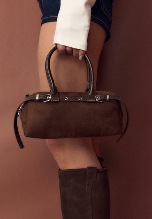 Roka drži majhno torbico iz temno rjavega semiša z dvojno ročico, oblečene v denim kratke hlače in temno rjave semiš čevlje, na rjavi podlagi.