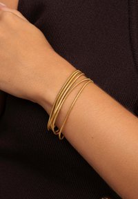 Bracciali in metallo color oro, quattro in totale, con una texture liscia e leggermente intrecciata, appoggiati su una pelle chiara contro uno sfondo di tessuto lavorato scuro.