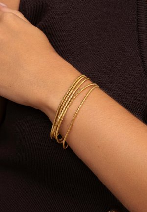 Brazaletes de metal en tono dorado, cuatro en total, con una textura lisa y ligeramente retorcida, descansando sobre un tono de piel claro contra un fondo de tejido oscuro.