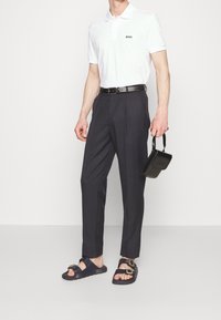 Vitt pikétröja med svart logotyp, marinhöga byxor, svart läderbälte och svarta sandaler med remmar. Håller en liten svart väska.