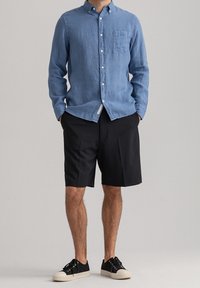 Camicia azzurra chiara con maniche lunghe e taschino sul petto, abbinata a pantaloni corti neri e sneakers nere con suole bianche.