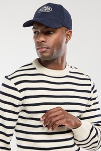 Granatowa czapka z sztruksu z zaokrąglonym rondem; sweter w paski w kolorze kremowym i granatowym z prążkowanymi mankietami i dekoltem. Zbliżenie na gest dłoni.