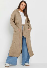 Beige lång cardigan med kapuschong, bälte i midjan, ribbade ärmslut och två framfickor, bärs över en vit topp och vida byxor.