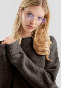 Okulary przeciwsłoneczne z złotą oprawą i fioletowymi soczewkami nosi osoba ubrana w grafitowy, teksturowany sweter z szerokimi mankietami.