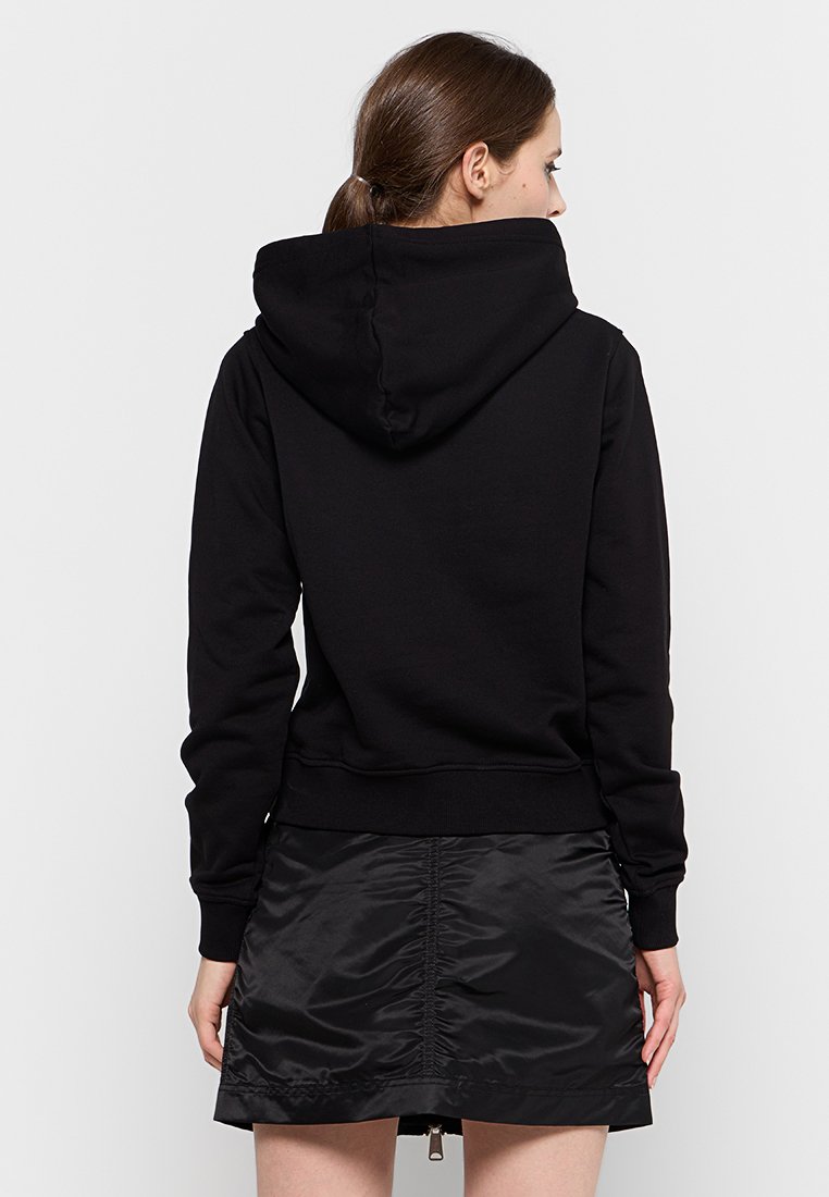 Haut à capuche noir avec une capuche spacieuse, des poignets et un ourlet côtelés. Texture lisse, coupe décontractée, associé à une jupe noire brillante.