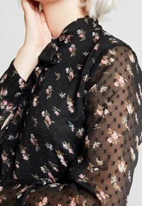 Svart blommig blus med långa, genomskinliga ärmar. Har en prickig textur, små blommönster i rosa och vitt samt en hög krage.