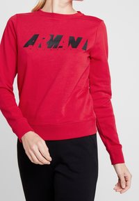 Felpa rossa con maniche lunghe, scollatura a girocollo e testo grafico nero "ARMANI EXCHANGE" sul davanti. Texture morbida e design aderente.