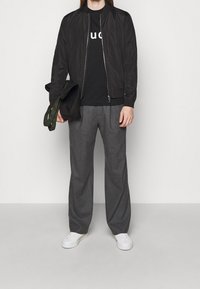 Personne portant une veste noire zippée, un t-shirt graphique noir, un pantalon large gris, des baskets blanches, tenant un sac fourre-tout noir avec des anses vertes.