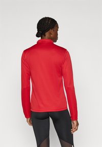 Κόκκινο αθλητικό τοπ με μακριά μανίκια και ψηλό γιακά, κατασκευασμένο από λείο ύφασμα. Διαθέτει εφαρμοστή σχεδίαση με αδιάκοπη πλάτη και μακριά μανίκια.