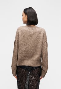 Καφέ μάλλινο πουλόβερ με ριμπ τελείωμα, χαλαρή εφαρμογή και πλαϊνά κουμπιά. Φοριέται πάνω από μαύρη φούστα με παγιέτες, παρουσιάζοντας αντίθεση υφής υφάσματος.