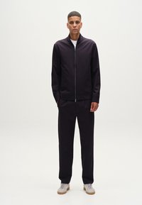 Chaqueta de zip oscuro en color púrpura con puños acanalados y pantalones a juego. Tejido texturizado, combinada con zapatillas de colores claros para crear contraste.