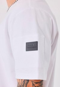 Primer plano de una camiseta blanca de manga corta con un parche rectangular negro etiquetado "PXP CONNEXION" en el brazo superior, por encima de un antebrazo tatuado.