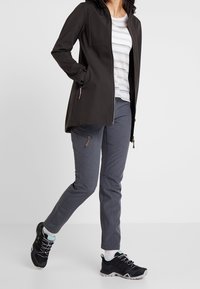 Chaqueta negra resistente al agua con cierre delantero, combinada con pantalones grises con bolsillos laterales y zapatillas deportivas negras con acentos en teal.