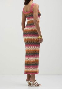 Robe sans manches en tricot avec un motif en zigzag orange, rose, blanc et vert foncé. Silhouette ajustée, longueur sous le genou, accompagnée de sandales à talons blanches.