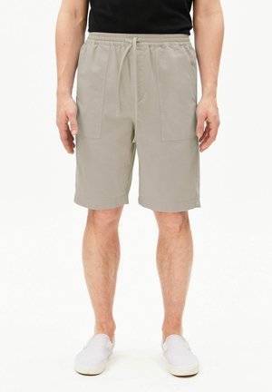 Mann trägt beige Shorts mit Kordelzug, schwarzes Hemd und weiße Slip-On-Schuhe, steht mit entspannten Armen an den Seiten vor weißem Hintergrund.
