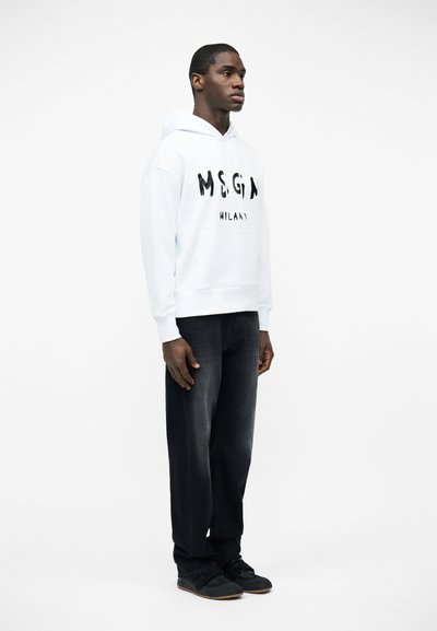 Άνδρας που φοράει λευκό hoodie MSGM Milano, μαύρο τζιν και μαύρα παπούτσια, στέκεται μπροστά σε ένα απλό λευκό φόντο.