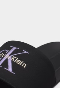 Mule noire Calvin Klein avec un "K" brodé en violet clair et le texte du logo "Calvin Klein" en blanc sur la bride.