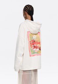 Sudadera crema con un gran gráfico colorido en la parte de atrás que presenta diseños florales y texto, además de capucha con cordón y un ajuste relajado.