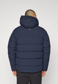 Mørkeblå jakke med hette, med en kviltet design, elastiske mansjetter og en opphengsløyfe bak. Teksturert stoff for isolasjon.