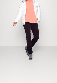 Vrouw die loopt in zwarte broek, koraalkleurige fleece met rits, witte pufferjack en grijze wandelschoenen tegen een effen lichte achtergrond.