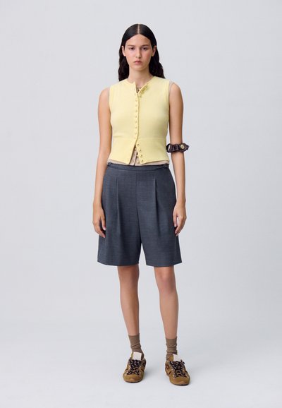 Cardigan senza maniche di un giallo chiaro con dettagli in bottone, abbinato a pantaloni corti grigi a pieghe e scarpe stringate marroni, accentuato da un bracciale marrone.
