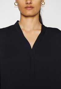 Zwarte blouse met een V-hals, voorzien van subtiele stiksels langs de randen. Glad stof met een zachte textuur. Geen zichtbare patronen of accenten.
