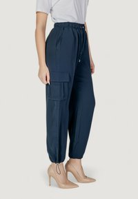 Pantaloni jogger navy con coulisse, dotati di tasca laterale cargo e polsini elastici, abbinati a tacchi alti color nude, su uno sfondo neutro.