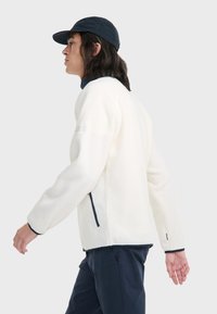 Weiße Fleecejacke mit hohem Kragen, marineblauen Akzenten, seitlichen Reißverschlusstaschen und langen Ärmeln. Getragen mit einer dunklen Mütze.